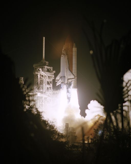Dramatic scene of Space Shuttle Atlantis lifting off at night for docking mission with Mir Space Station in 1997. Featuring fiery rocket launch framed by Florida foliage, illuminating night sky. Useful for topics on space missions, aerospace achievements, NASA history, and technology advancements.