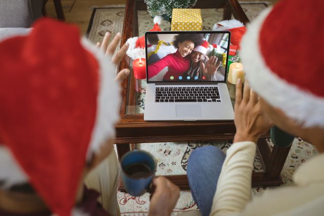 Couple wearing Santa hats using a laptop for a video call with African American friends, who are on the screen, also wearing festive hats. Ideal for use in articles or advertisements focusing on modern holiday celebrations, remote connections during festive seasons, virtual greetings, and the use of technology to stay connected with loved ones.