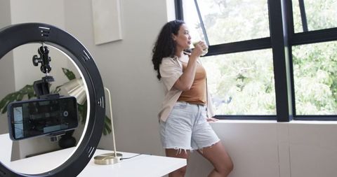 African american woman standing by window sipping from cup while recording with ring light