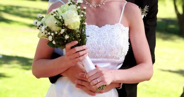 Newlywed couple embracing with bride holding bouquet of white roses and delicate flowers. Captures intimate moment with focus on bridal attire and floral arrangement. Suitable for wedding industry promotions, romantic event invitations, or marriage-related content.