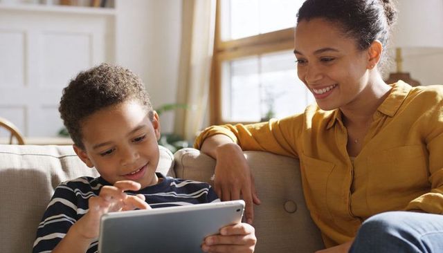 Mother and son learning together on tablet smiling in sunlit cozy living room