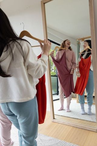Diverse friends laughing while holding dresses and heels by mirror in cozy loungewear