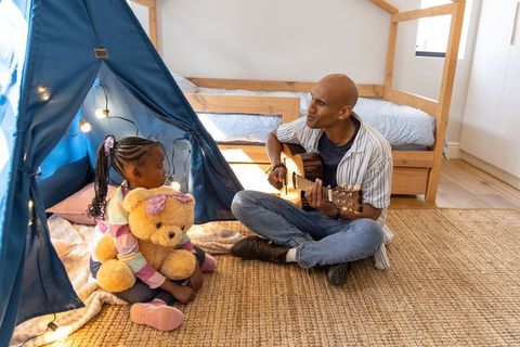 African american father playing guitar while daughter hugs teddy in sunlit bedroom teepee