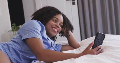 Relaxed african american woman lying in satin pajamas smiling while checking smartphone
