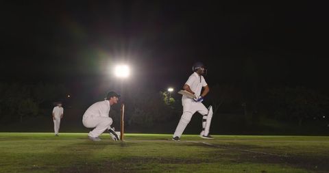 Night cricket match under floodlights with batsman running and wicketkeeper crouching