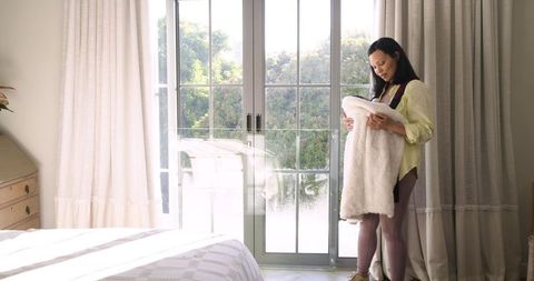 Middle-aged Asian Woman Holding Plush Blanket in Sunlit Bedroom