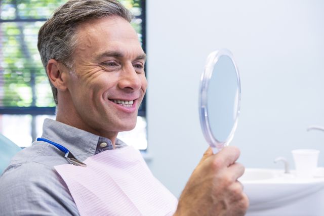 Middle-aged man smiling while looking in mirror at dental clinic. Ideal for use in advertisements for dental services, oral health campaigns, and healthcare promotions. Highlights patient satisfaction and positive dental experiences.