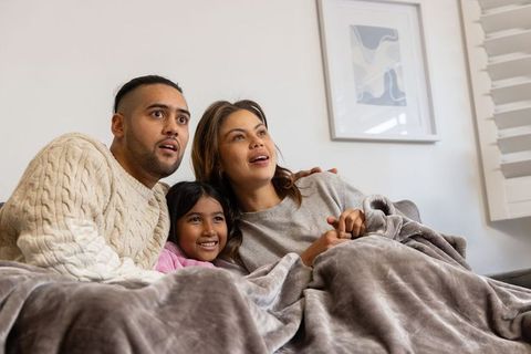Family cuddling on couch under plush blanket watching movie together