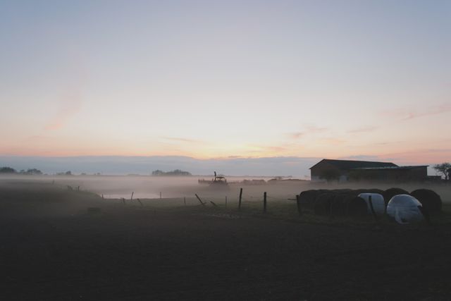 The image features a peaceful rural landscape at dawn with a light mist covering the ground and a barn in the background. Hay bales are visible, and the sunrise casts a soft light over the scene. Ideal for themes related to agriculture, farming, tranquility, and the beauty of rural life.
