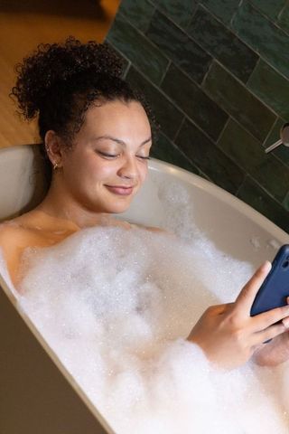 African american woman relaxing in bathtub with bubbles using smartphone in bathroom