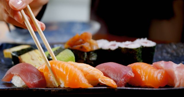 Close-up of a hand holding chopsticks, picking up assorted sushi pieces including salmon and tuna from a plate. Perfect for illustrating concepts related to Japanese cuisine, dining experiences, sushi making, and food presentations.