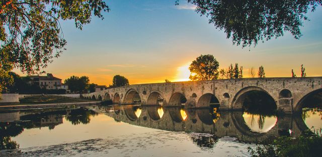 Vintage stone bridge reflecting on calm river during sunrise. Ideal for travel blogs, nature photography, historical sites. Suitable for backgrounds, website banners, postcards, and tourism material, emphasizing tranquility and beauty of nature.