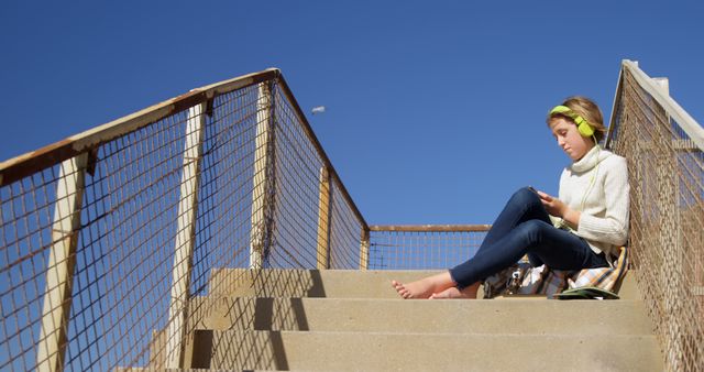 Happy caucasian girl in sweater sitting on stairs and using headphones and smartphone on sunny day. Childhood, communication, free time, summer, travel, vacations and lifestyle, unaltered.