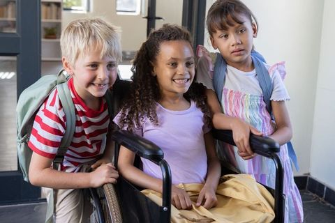 Multiethnic school friends supporting classmate in wheelchair smiling at school entrance