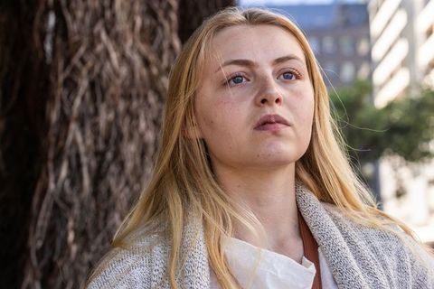Young woman standing by tree on city sidewalk wearing knit cardigan looking thoughtful