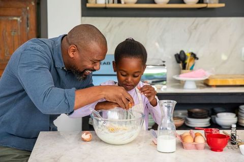 African american father guiding daughter cracking eggs, mixing batter on marble countertop