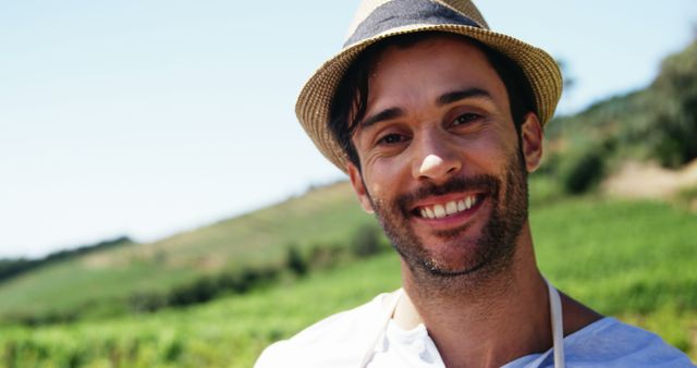 Smiling man with beard wearing a hat and apron standing outside in bright, sunny countryside with sprawling greenery. Suitable for content about outdoors, happy mood, summer day, and rural lifestyle. Can be used in articles on leisure activities, fashion blogs highlighting casual wear, or marketing for outdoor events and travel.