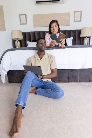 Diverse couple planning international travel comparing passports with tablet on bed