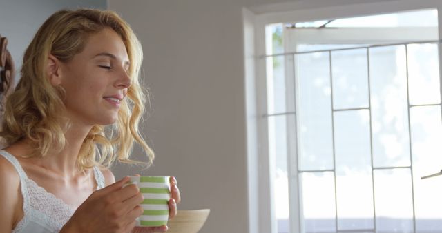Young blonde woman sipping coffee in a sunny room, creating a perfect depiction of morning relaxation and contemplation. Ideal for lifestyle, health, and wellness themes, promotions for coffee or tea products, and articles about self-care or mindfulness.