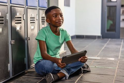 African american boy using tablet sitting by school lockers for digital learning