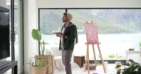 Indian artist studying pink abstract on easel in modern coastal studio with tablet