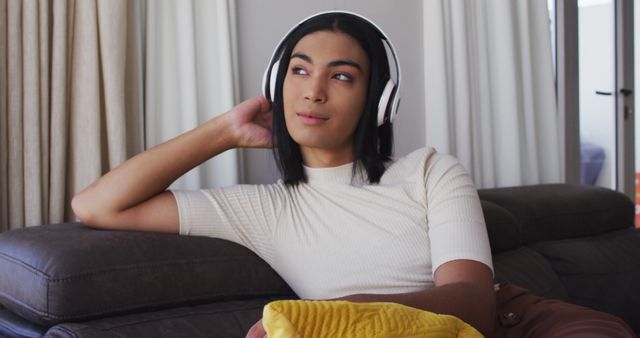 Gender fluid male wearing headphones listening to music while sitting on the couch at home. concept of gender expression, identity and diversity