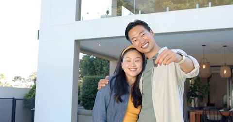 Asian couple celebrating new home with keys at modern two-story house, smiling homeowners