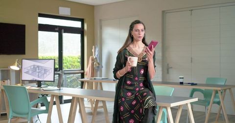 Indian woman in kurta sitting on table holding smartphone and coffee in minimalist studio