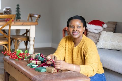 African american woman wrapping holiday gifts at home wearing cozy mustard sweater, festive decor