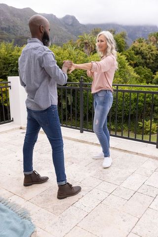 Diverse senior couple dancing on outdoor terrace holding hands with mountain view and greenery