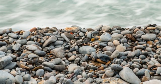 Scene of smooth pebbles on a beach shore with soft, gentle waves in the background. Suitable for use in projects related to nature, tranquility, coastal living, summer vacation, or as a natural background for various creative projects.