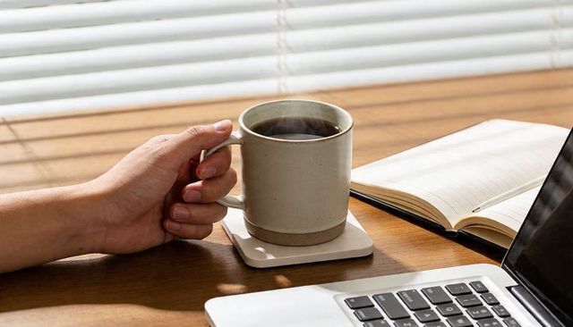 Holding coffee mug on wooden desk, cradling steaming cup near laptop and notebook