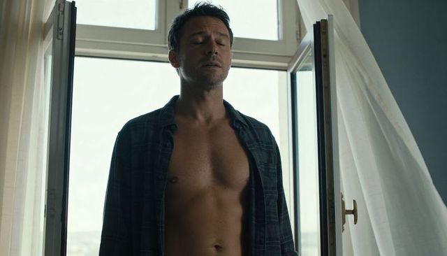 Shirtless man standing at open window, eyes closed, basking in soft morning balcony light