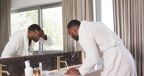 Morning grooming ritual: mature man in robe washing face at modern bathroom vanity