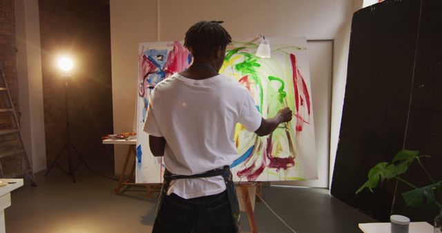 Young artist is seen from back while painting a colorful abstract artwork on canvas in a studio. Ideal for themes of creativity, modern art, and artistic activities. Useful for illustrating topics about young artists, abstract art techniques, and creative processes.