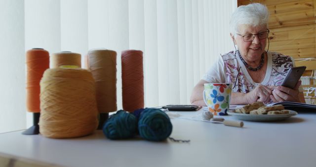 Senior Caucasian woman smiles while using a tablet at home, with copy space. She's surrounded by yarn and cookies, suggesting a cozy crafting break.