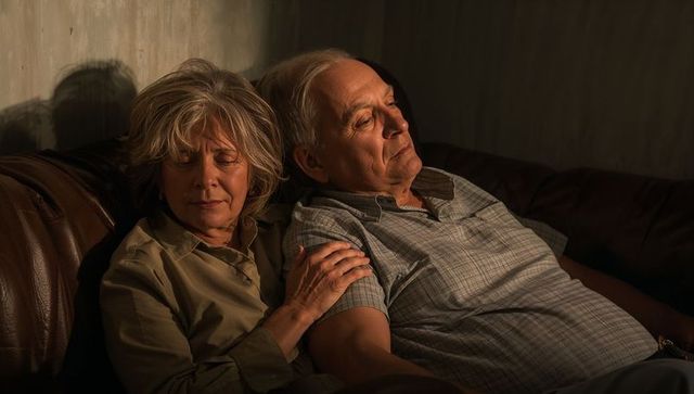 Older couple relaxing on leather sofa, woman holding partner's arm during quiet evening