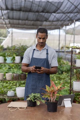 Asian man checking smartphone while managing greenhouse plants and sales at nursery