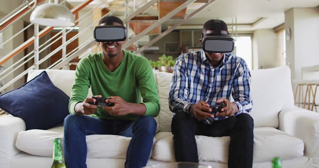 African american teenage twin brothers on couch using vr headsets and playing computer game smiling. family leisure time at home together during quarantine lockdown.