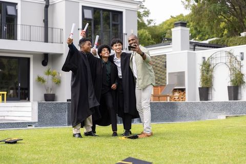 Diverse male family celebrating graduation on lawn posing for selfie with diplomas