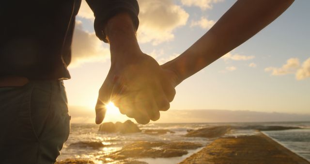 Couple holding hands at sunset by the sea, with copy space. Their romantic moment is enhanced by the warm glow of the setting sun.