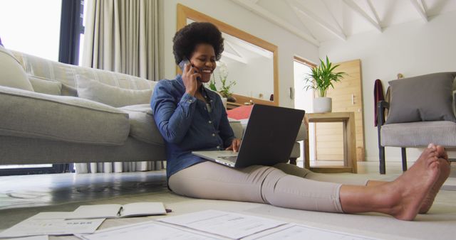 African American woman sitting on floor against sofa, working from home on a laptop while talking on phone. Scattered documents indicate busy work environment. Perfect for illustrating the modern home office, remote work lifestyle, or freelance work settings.