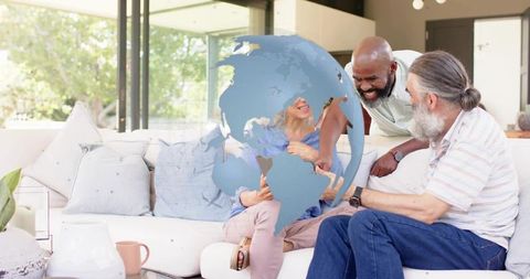 Diverse friends sitting on sofa pointing at digital globe overlay in modern living room