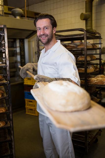 Professional baker smiling while holding freshly baked bread in a commercial kitchen. Ideal for use in content related to bakery businesses, culinary arts, artisan baking, and cooking classes. Perfect for illustrating the joy and skill of professional bakers and promoting products or services in the food industry.