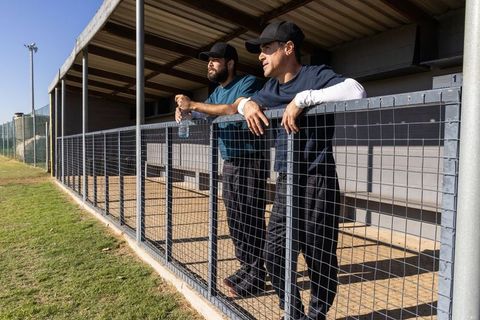 Baseball coaches leaning on dugout fence watching game on sunny field with water bottle