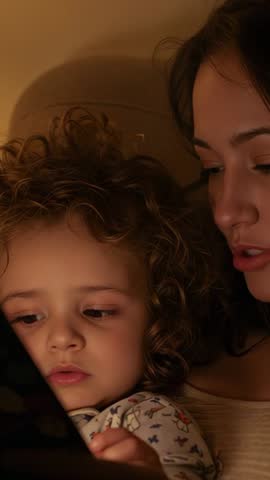 Vertical video: Mother narrating bedtime story while toddler taps tablet, cuddling close