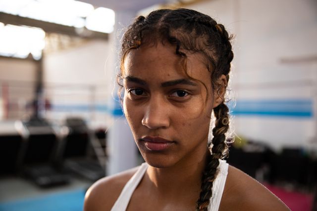 This image captures a confident biracial female boxer in a gym, wearing sports clothes and looking directly at the camera with a focused expression. Ideal for use in articles or advertisements related to sports, fitness, women's empowerment, athletic training, and motivational content.