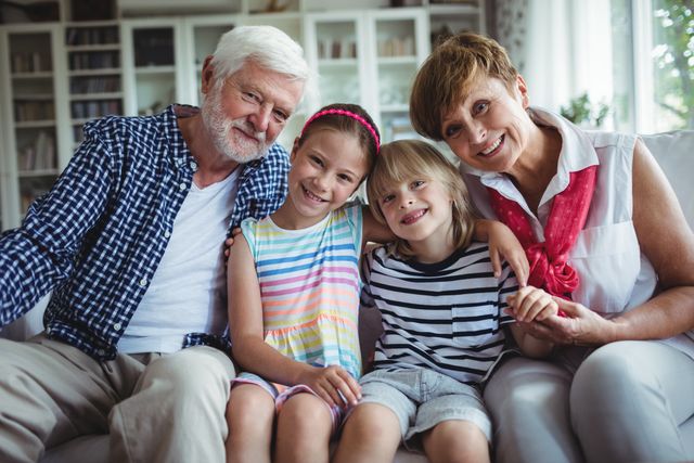 Grandparents and their grandchildren are sitting together on a couch in a cozy living room. They are smiling and showing affection, highlighting the bond and love within a multigenerational family. This image is perfect for use in family-oriented advertisements, articles about family dynamics, or promotional materials for family services.