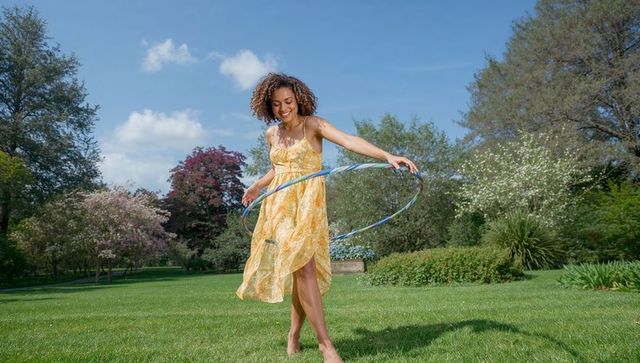 Joyful woman hula hooping in yellow floral sundress on sunlit park lawn curly hair barefoot