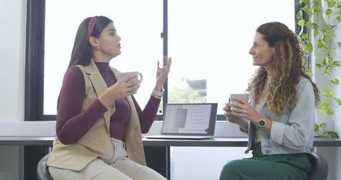 Female coworkers sharing ideas over coffee in modern bright office with laptop and plants